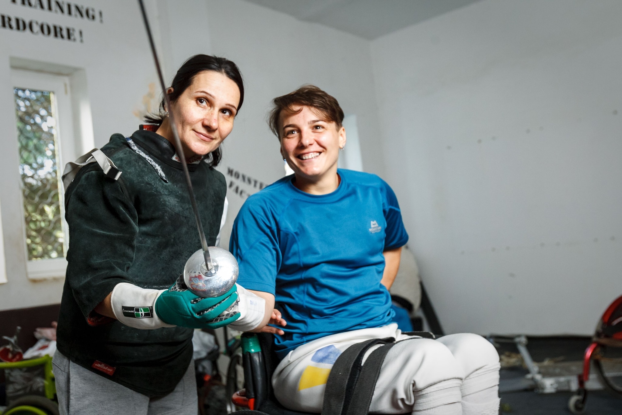 A coach in fencing kit smiling beside a woman in a sports wheelchair holding an epee