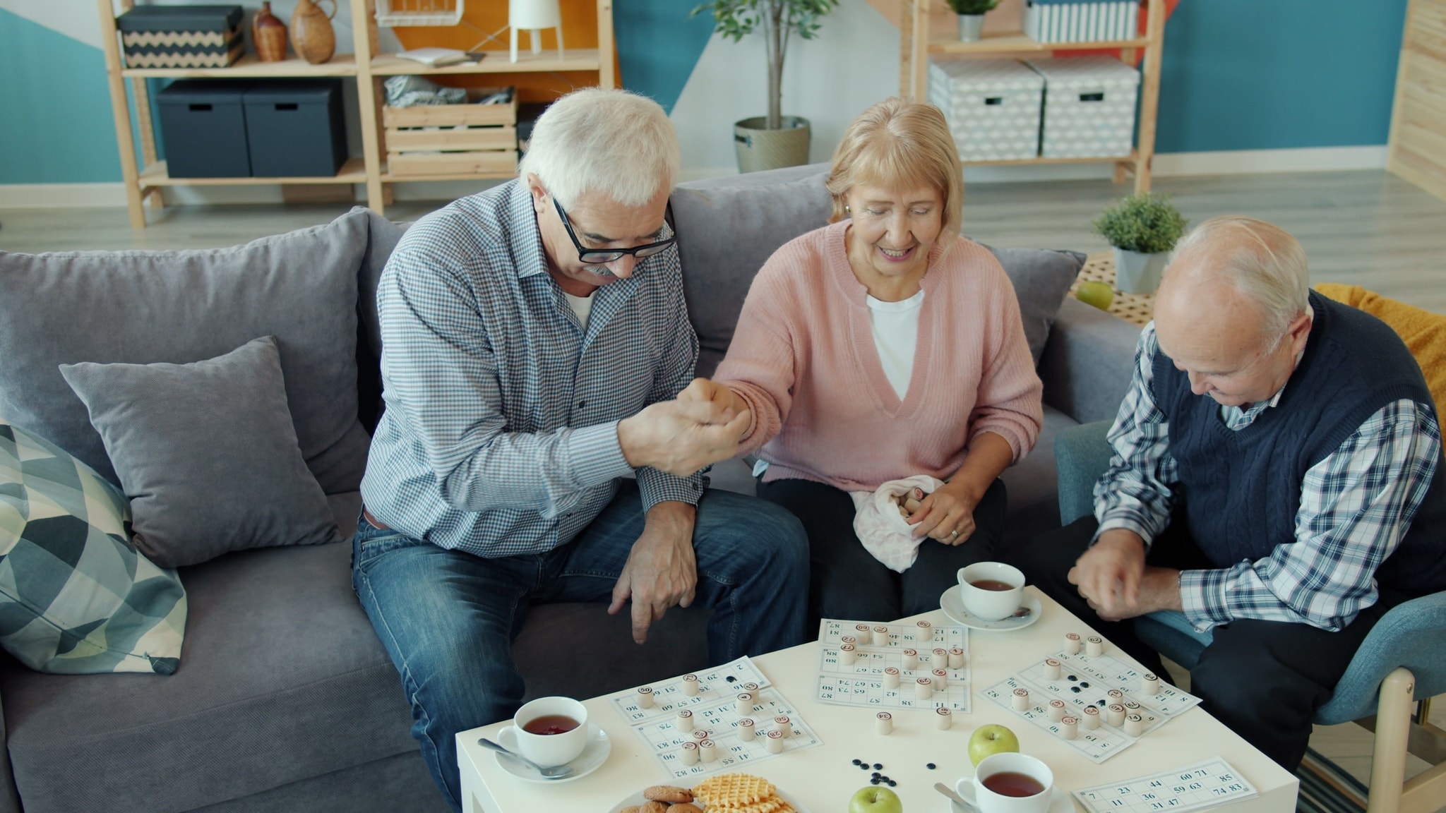 Two older people laughing together over a game of bingo, tea on the table